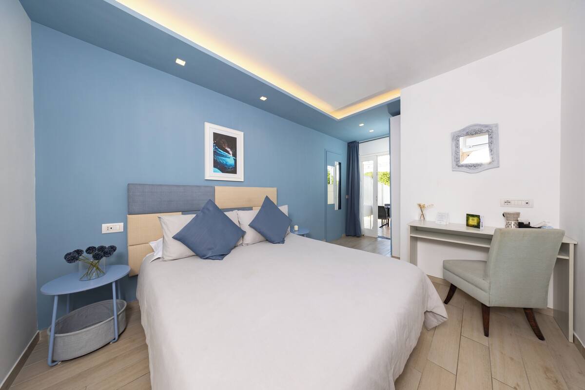 A bedroom with a bed, a desk, a chair, a small table with a vase, and a wall-mounted mirror.