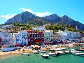 Un pueblo costero con edificios de colores, un puerto deportivo con barcos y montañas al fondo.