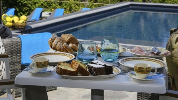Una mesa con alimentos para desayuno cerca de una piscina, incluyendo croissants, pan y pastel, con vistas a la piscina y vegetación al fondo.