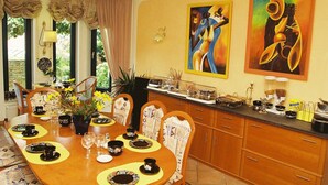 A dining area with a wooden table set for four, surrounded by chairs. There are two paintings on the wall, a buffet counter with various food items, and a window with curtains. - Hotel Garni Schneevoigt (Hagenburg)