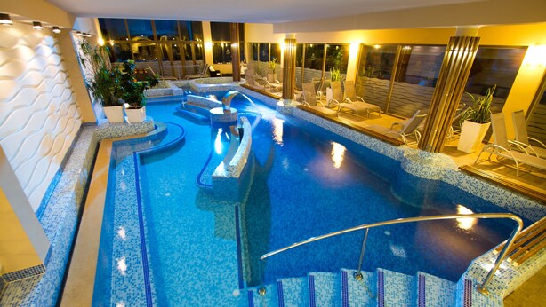 A large indoor swimming pool with multiple lanes, a seating area with chairs, and a staircase leading down to the pool. - Piknik Wellness Hotel (Siófok)