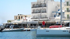 Un port de plaisance avec des bateaux amarrés, une personne debout sur un bateau, et des bâtiments avec des enseignes en arrière-plan.