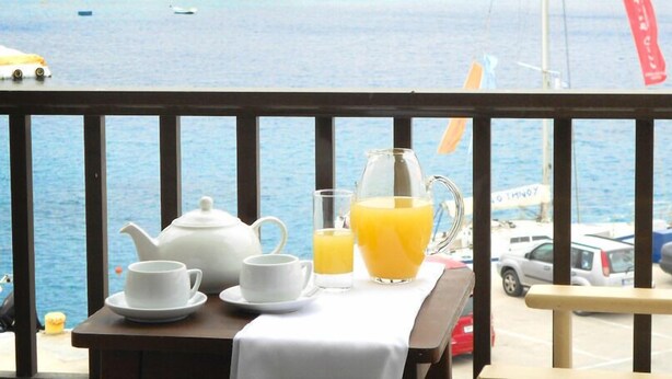 Une table avec une théière, des tasses et un verre de jus d’orange, placée sur un balcon donnant sur la mer.