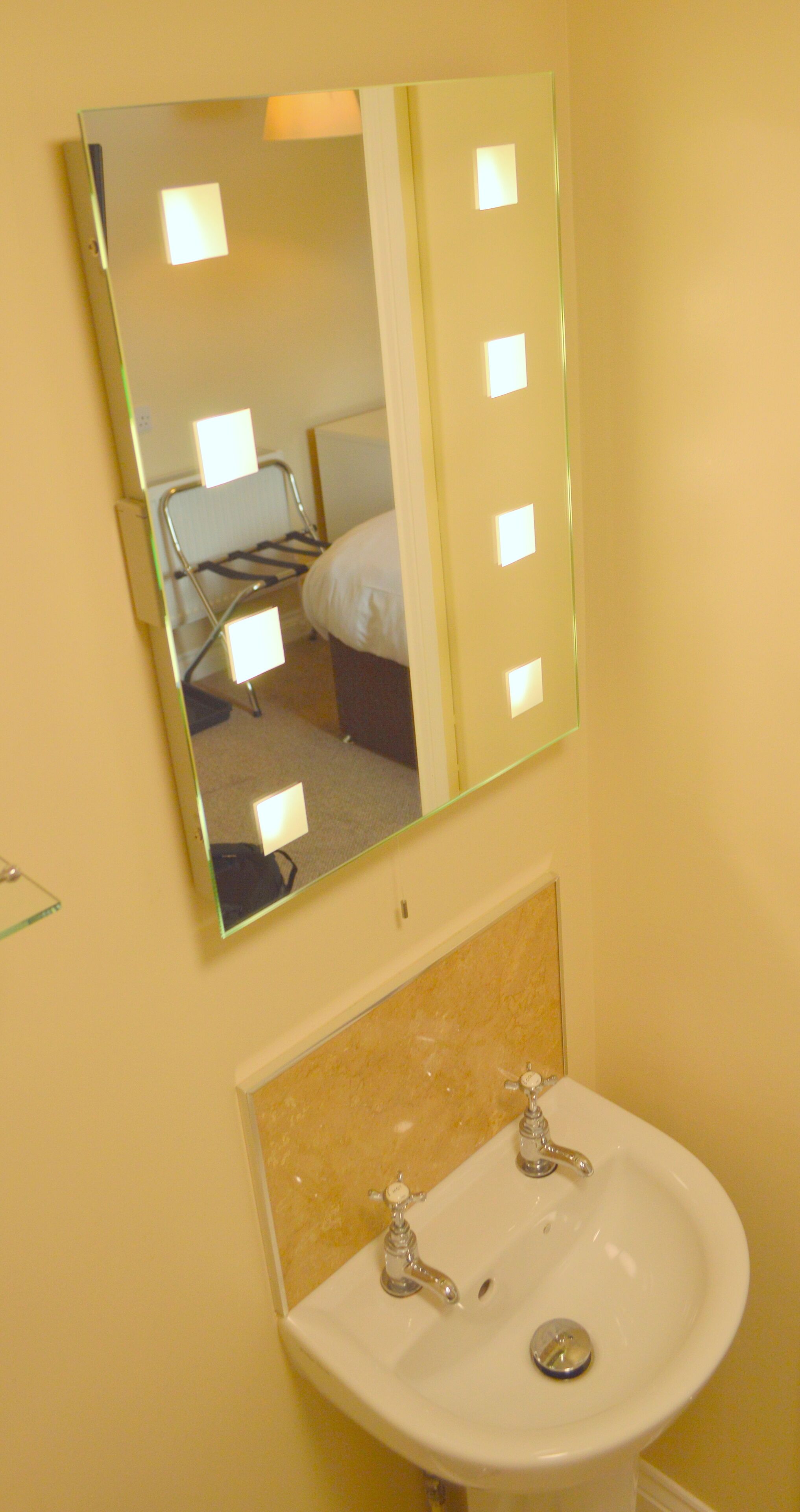 Casa de banho com lavatório, espelho e uma cama visível no reflexo.
