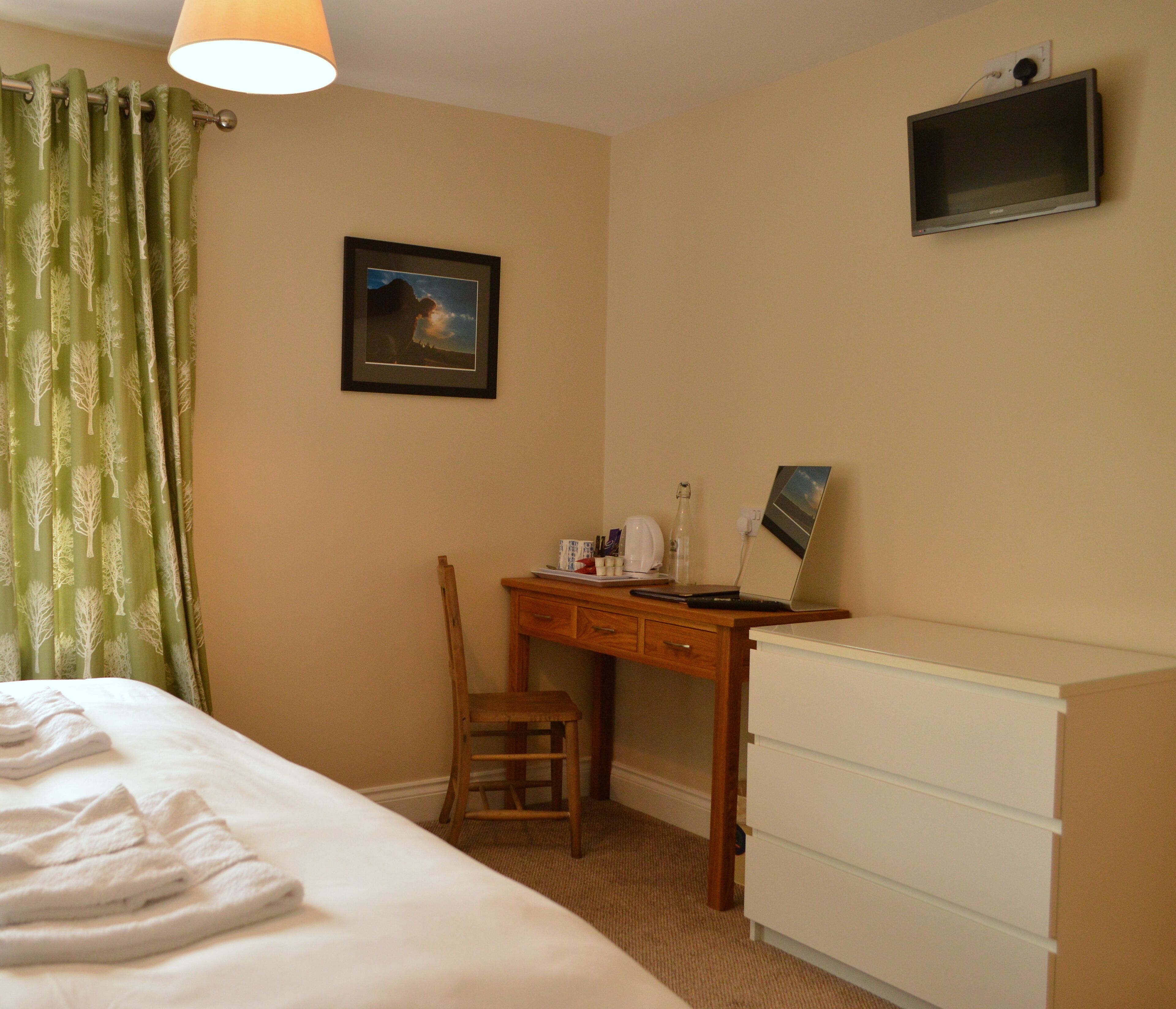 A hotel room with a bed, a desk with a chair, a dresser, a framed picture on the wall, and a television mounted above the dresser.