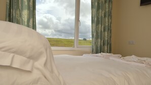 A neatly made bed with white linens, a window with curtains, and a view of a grassy field.