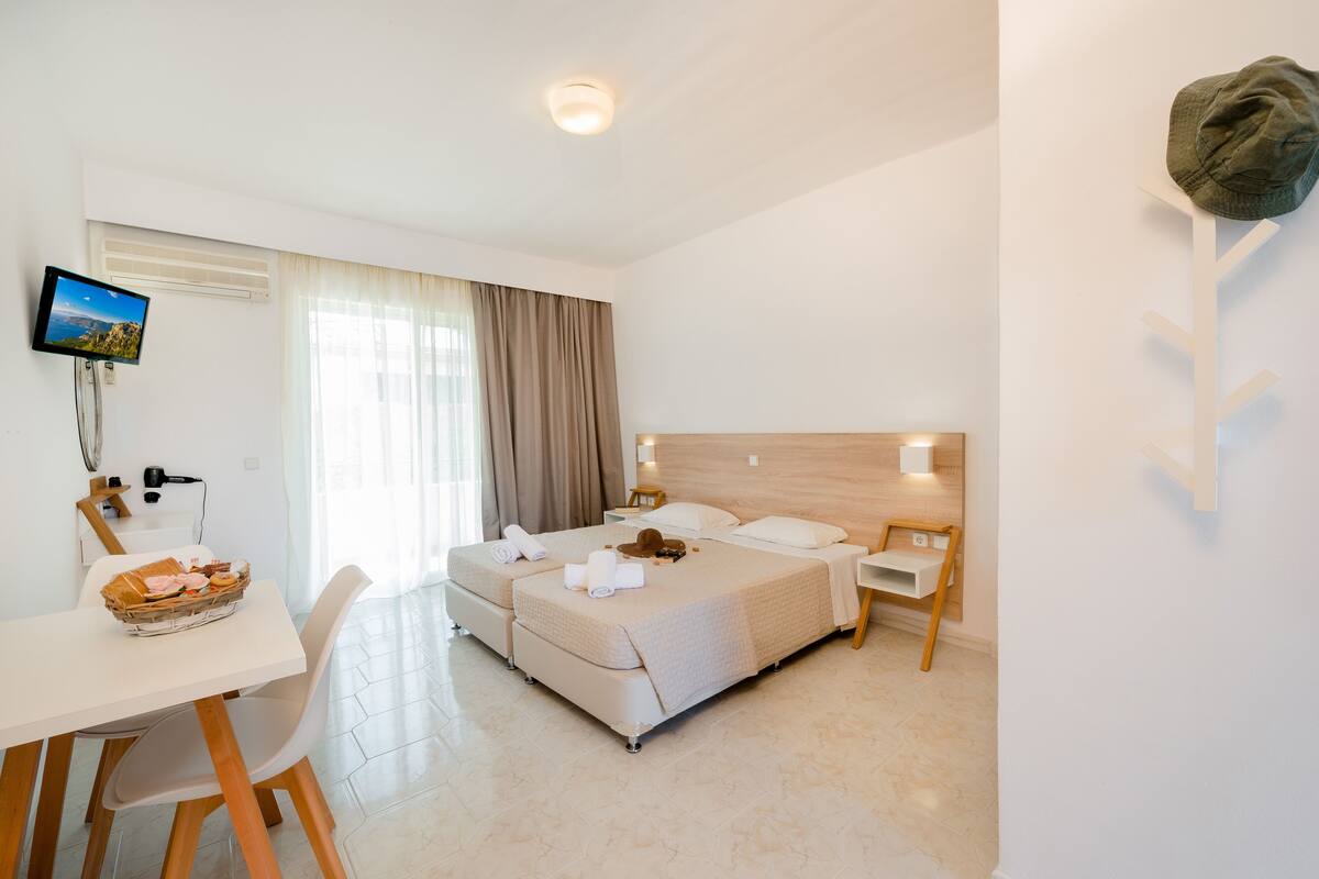 A hotel room with two beds, a desk with a basket of pastries, a TV, and a wall-mounted hat rack.