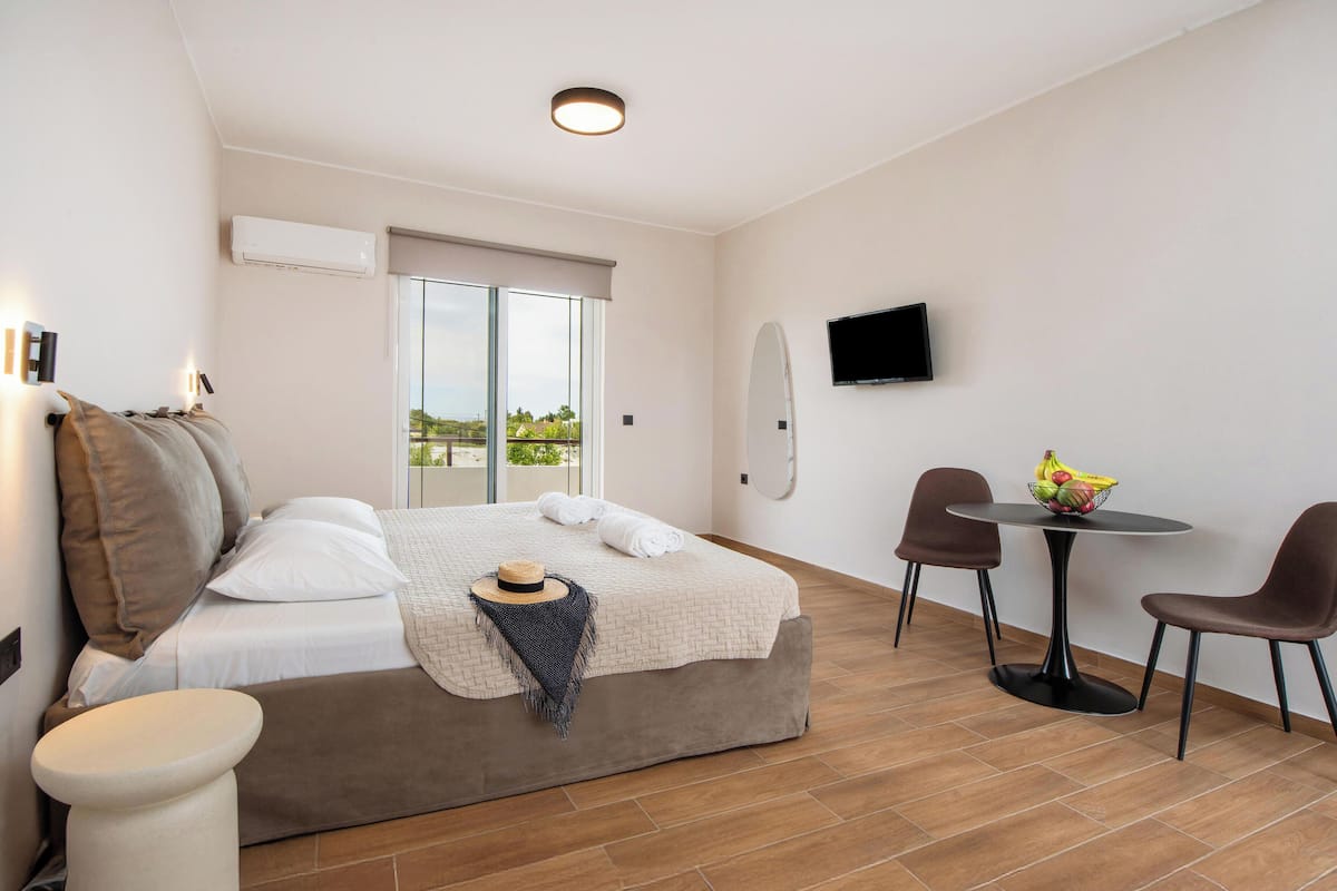 A modern hotel room with a bed, a small table with chairs, a TV, and a balcony with a view.