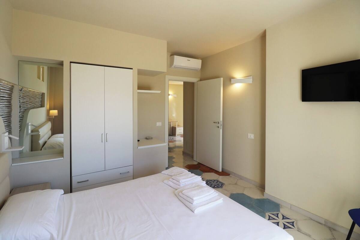 A hotel room with a bed, a wardrobe, a mirror, a television, and a hallway leading to another room.