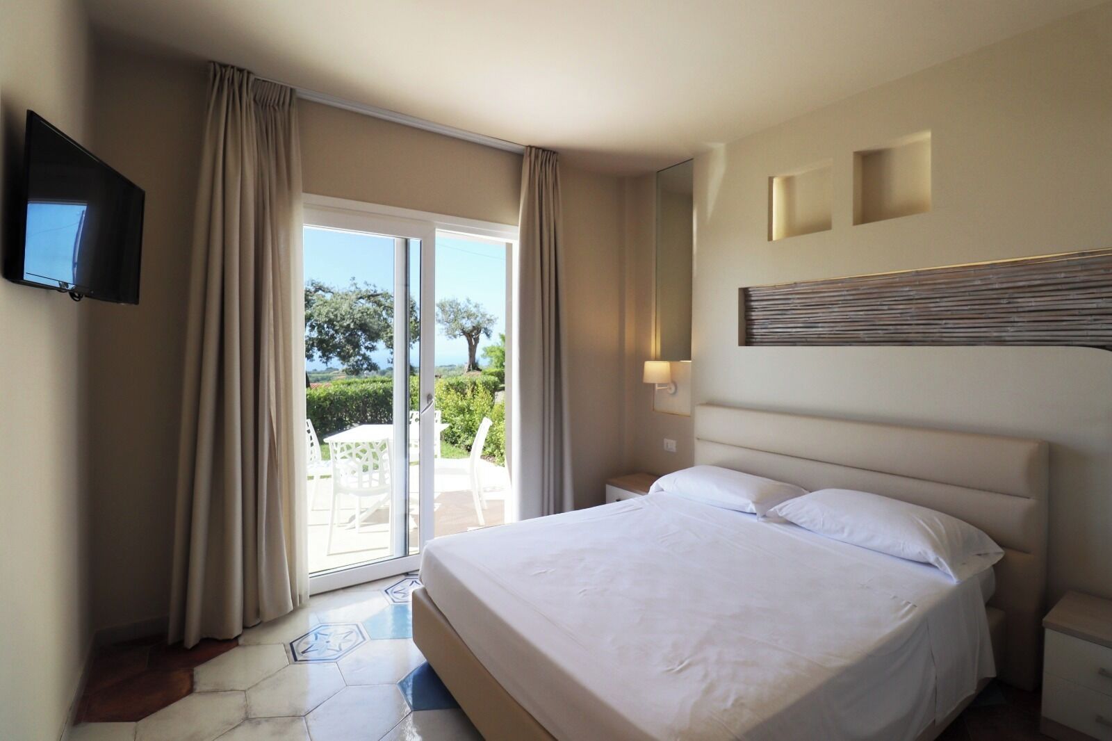 A hotel room with a bed, a TV, a sliding glass door to the outside, and a small table with chairs.