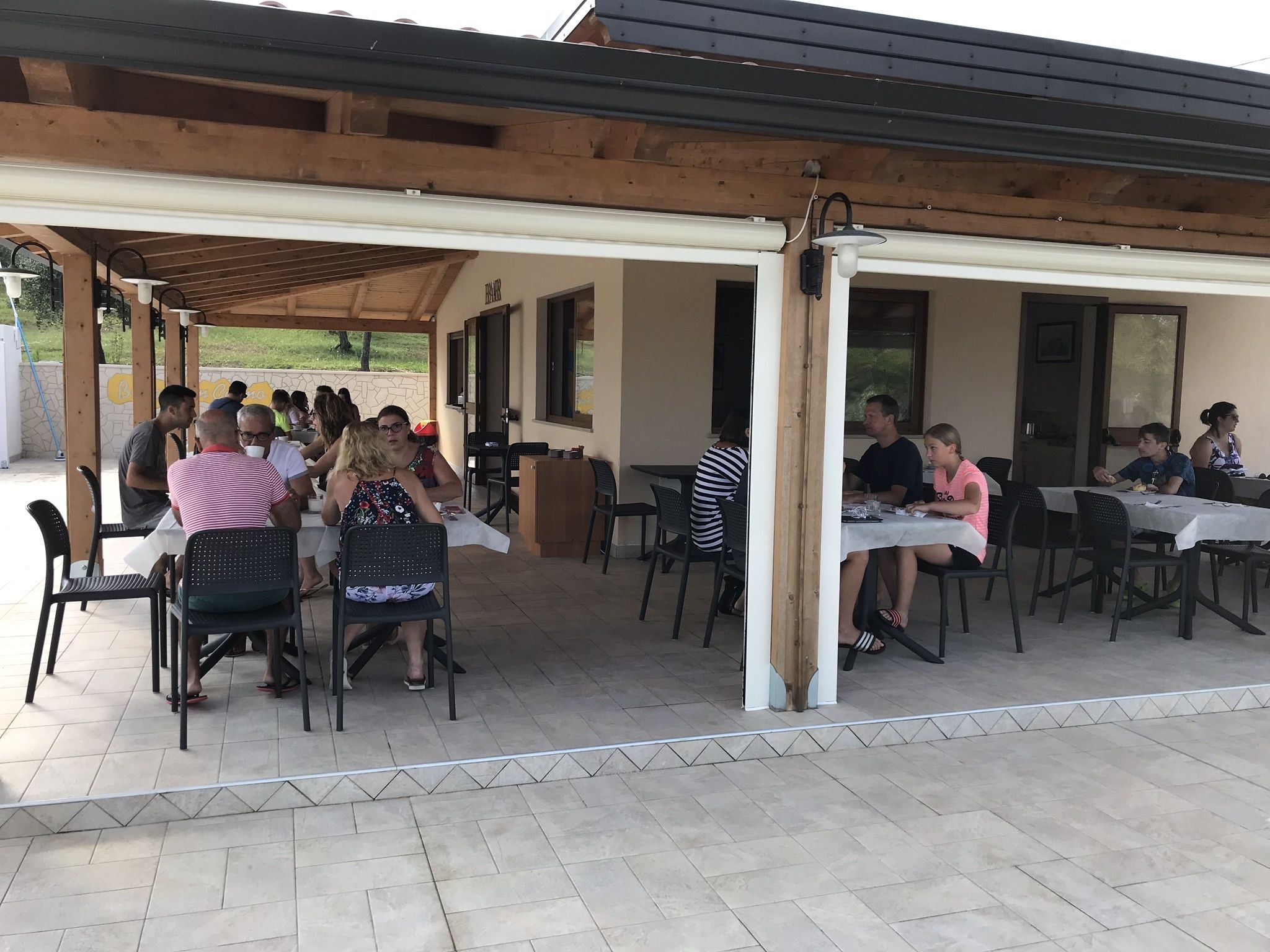 Een terras buiten met tafels en stoelen, overdekt door een houten constructie met een betegelde vloer.