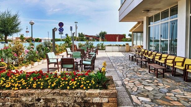 Un área de estar al aire libre con suelo de piedra y sillas, rodeada de macizos de flores y un edificio con grandes ventanales.