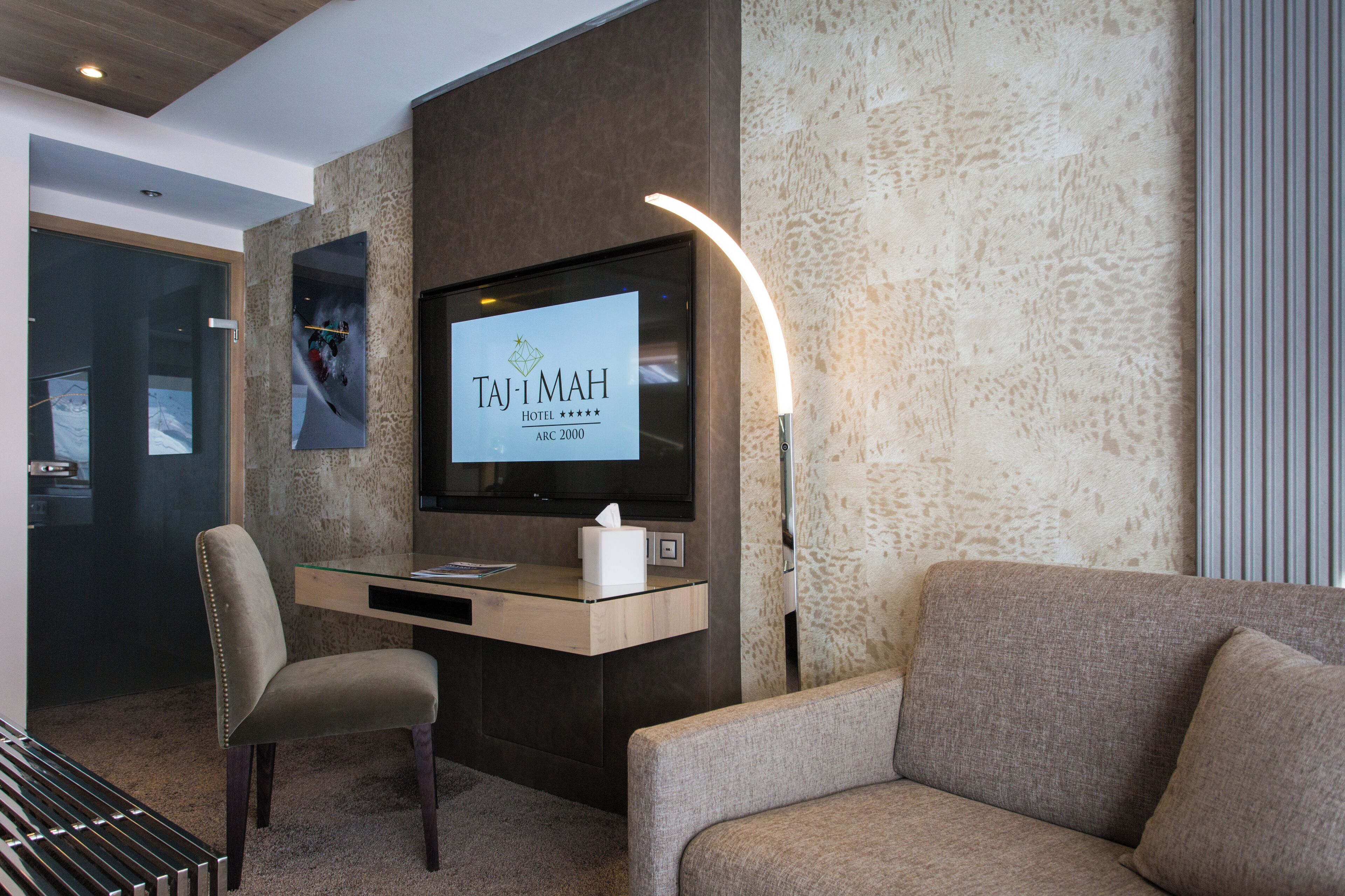 A hotel reception area with a flat-screen TV displaying the hotel name, a desk with a tissue box, a chair, and a sofa.