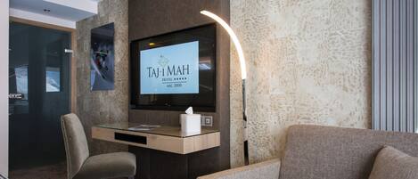 A hotel reception area with a flat-screen TV displaying the hotel name, a desk with a tissue box, a chair, and a sofa.
