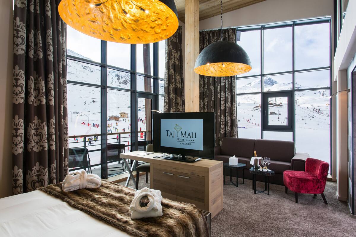 Hotellihuone, jossa on sänky, työpöytä, televisio ja näkymä lumiselle vuorelle.