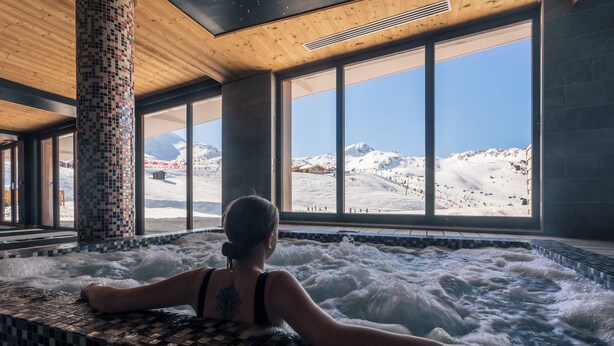 A person relaxing in a hot tub with a view of a snowy mountain landscape through large windows. - Hôtel Taj-I Mah by Les Etincelles  (Bourg-Saint-Maurice)