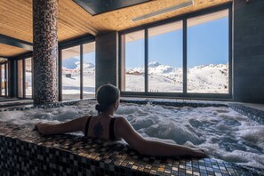 A person relaxing in a hot tub with a view of a snowy mountain landscape through large windows.