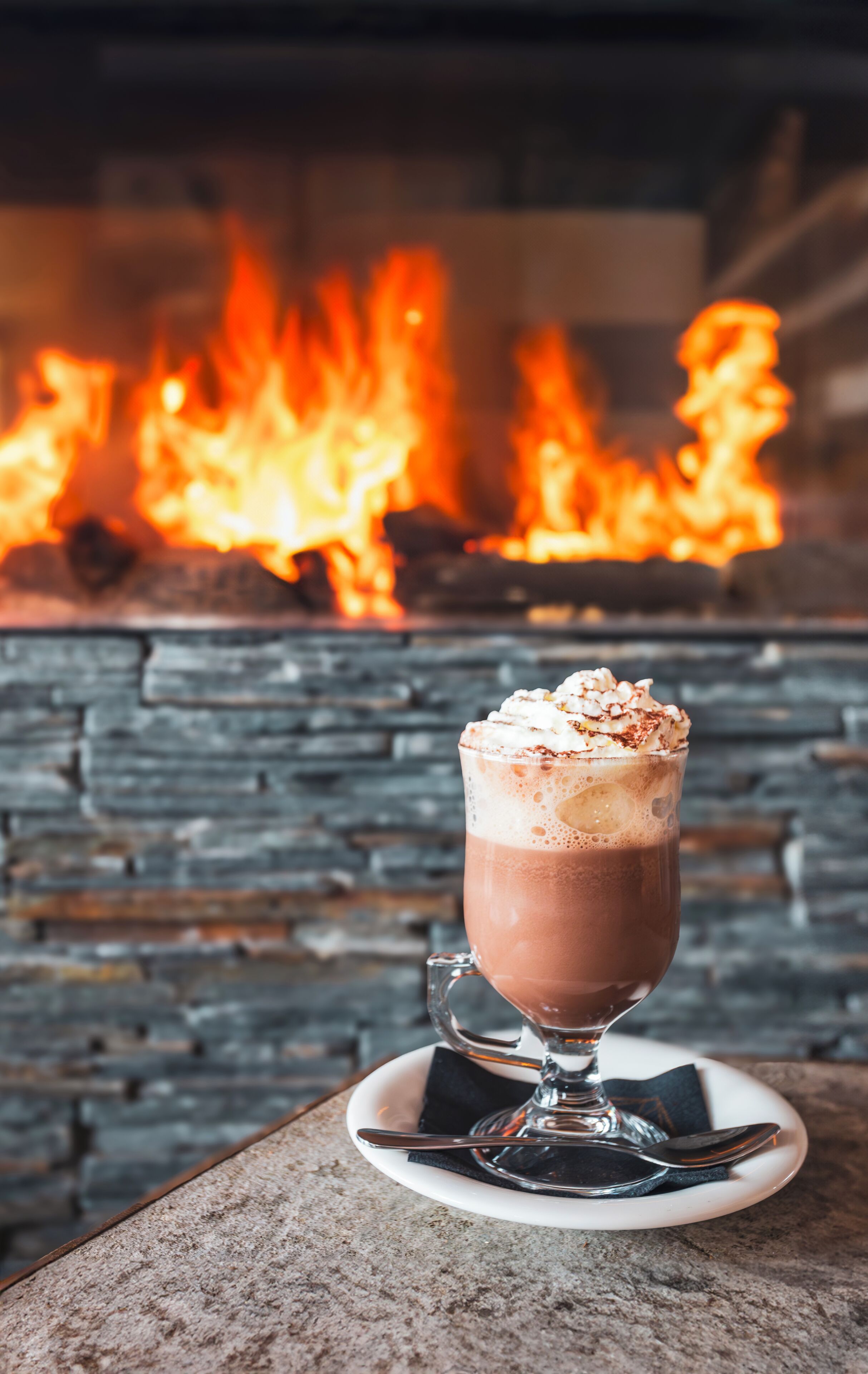 Una taza de chocolate caliente con crema batida sobre un posavasos frente a una chimenea con llamas.