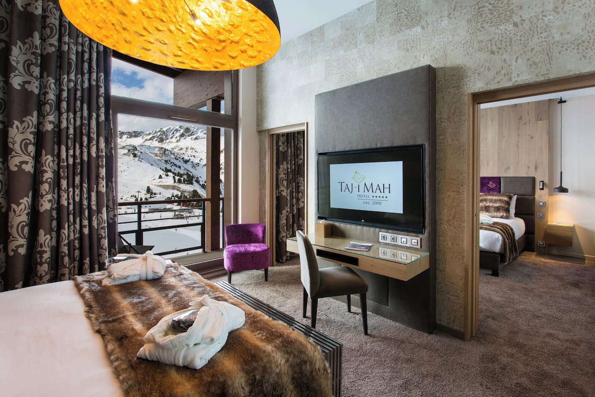 Hotellihuone, jossa on suuri sänky, työpöytä ja televisio, sekä näköala vuorille ja parveke.
