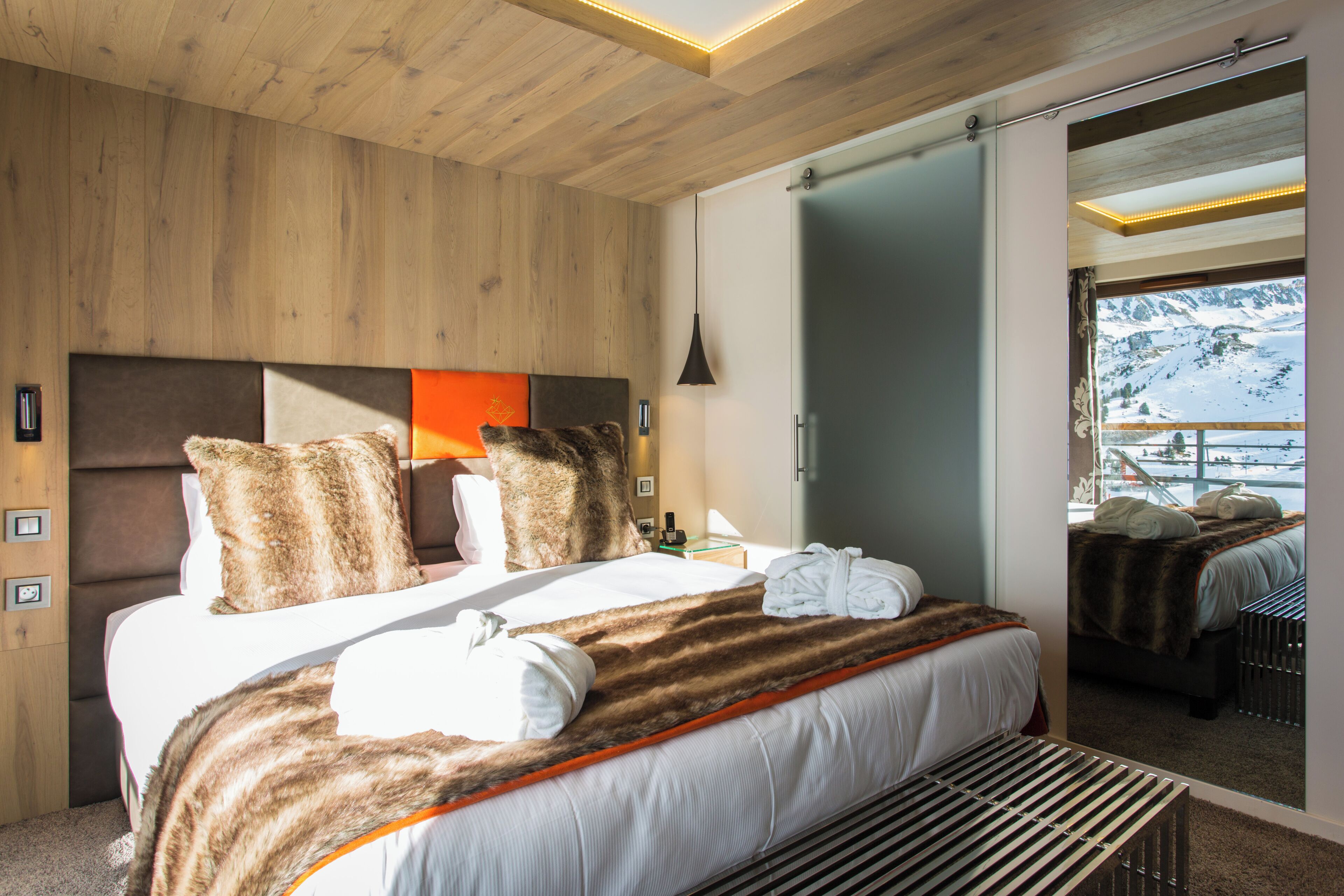 A modern hotel room with a large bed, wooden panel walls, a view of a snowy mountain, and a sliding door to a balcony.