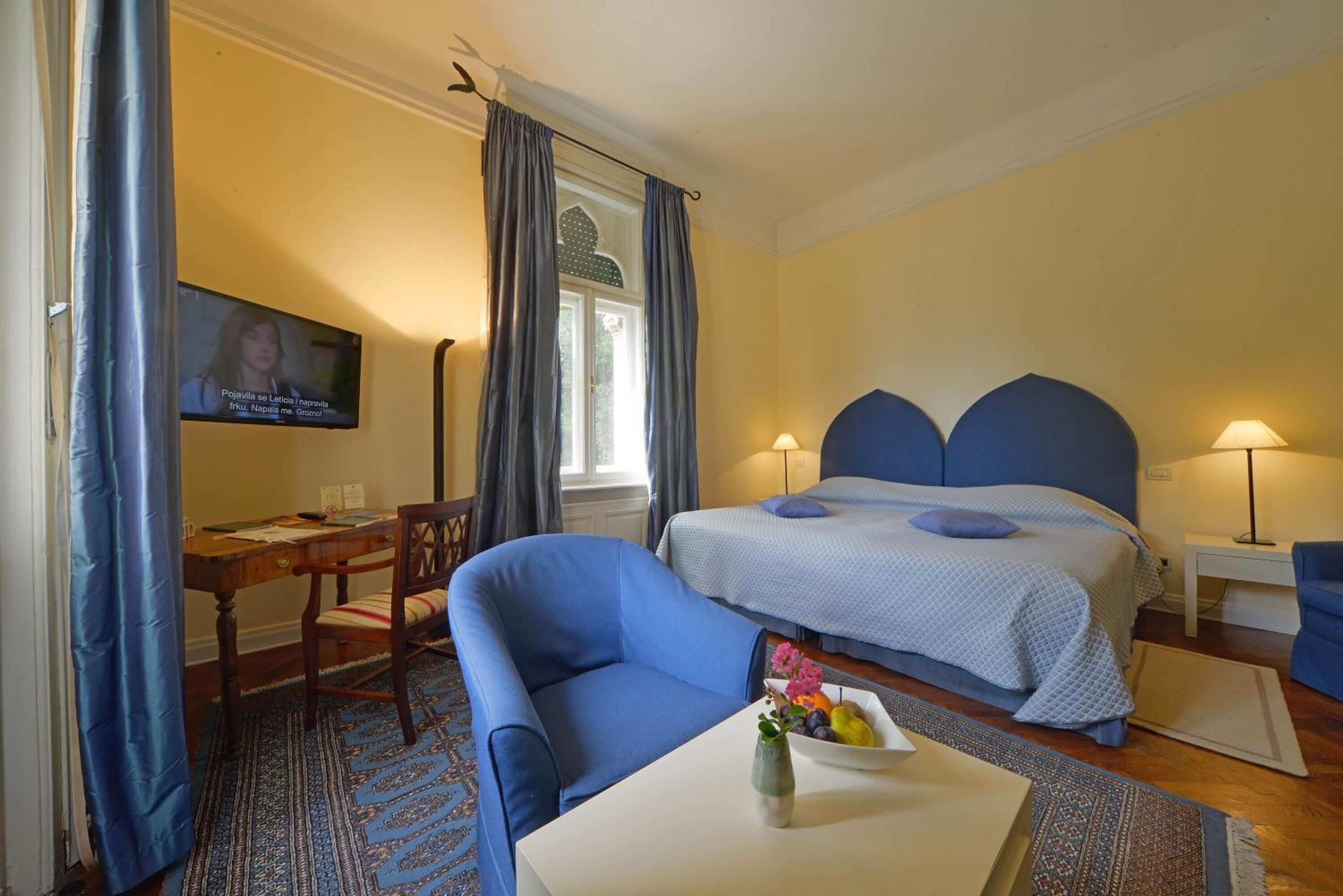 Une chambre d’hôtel avec un lit, un bureau, une chaise, une télévision et une petite table sur laquelle est posé un vase de fleurs.