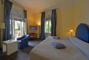 Habitación de hotel con cama, una silla, una mesita, televisión y ventana con cortinas.