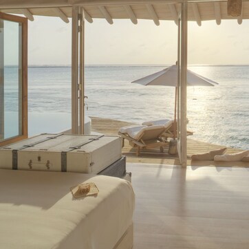 A room with a bed, a wooden floor, a view of the sea, and a beach with umbrellas.