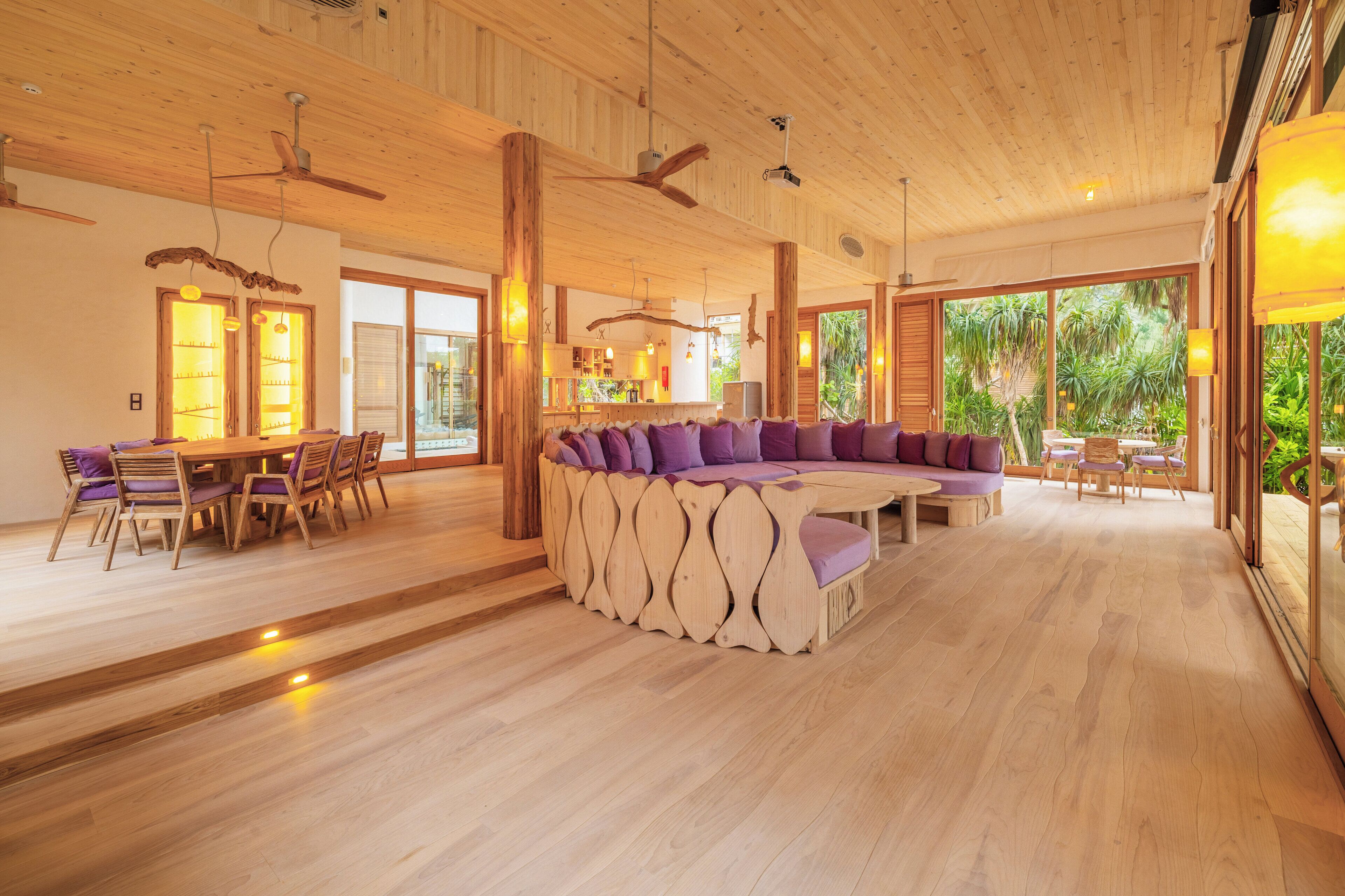 A spacious living area with a wooden ceiling, large windows, and a central seating area with purple cushions.