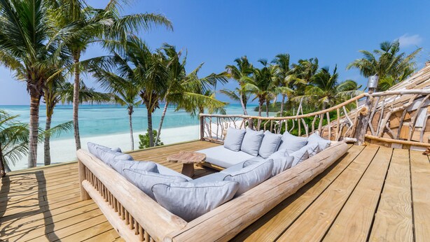 A wooden deck with a sofa and a table, overlooking a beach with palm trees and the ocean. - Soneva Jani (Medhufaru)