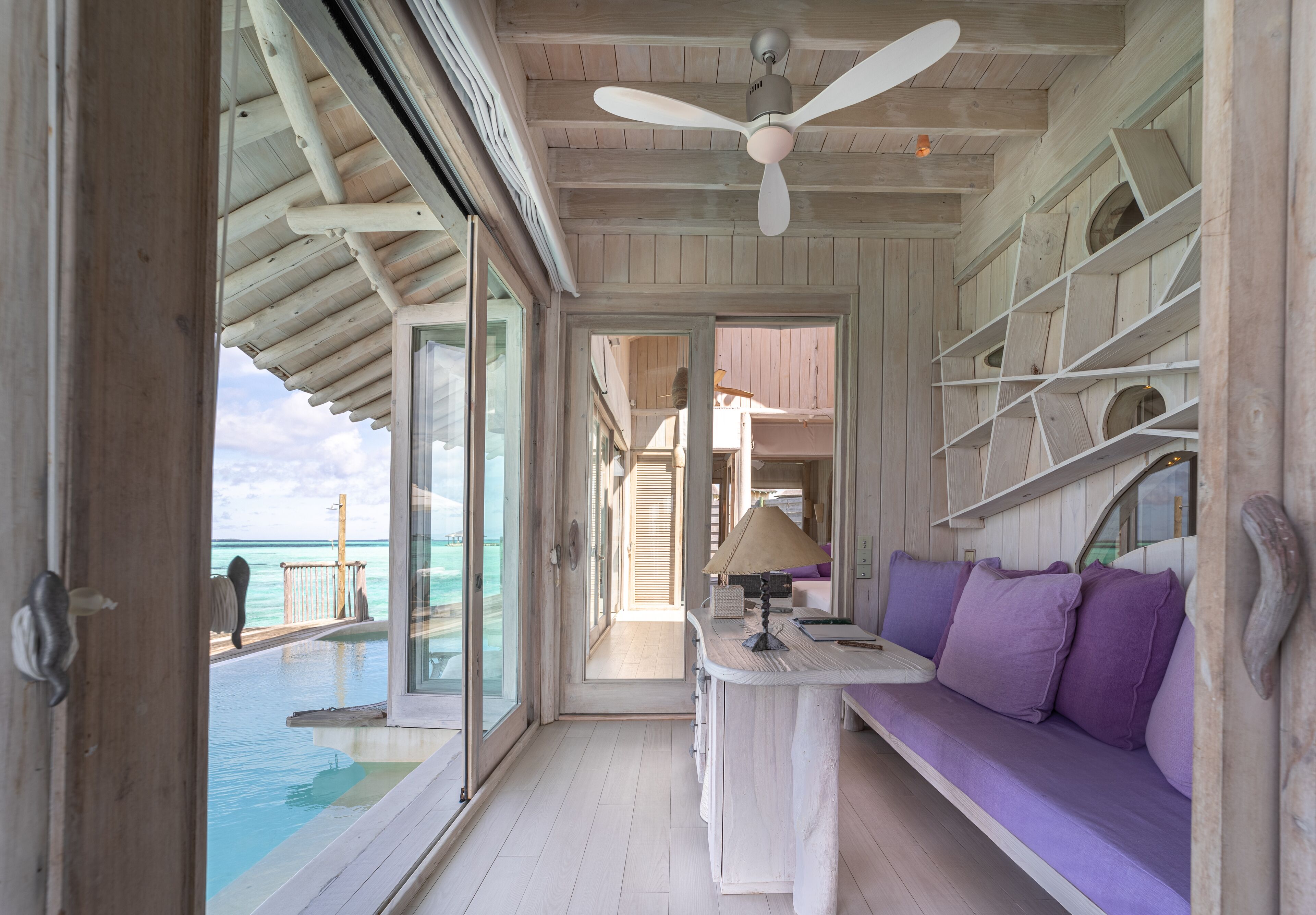 A room with a view of the ocean, a ceiling fan, a sofa with cushions, and a table with a lamp.