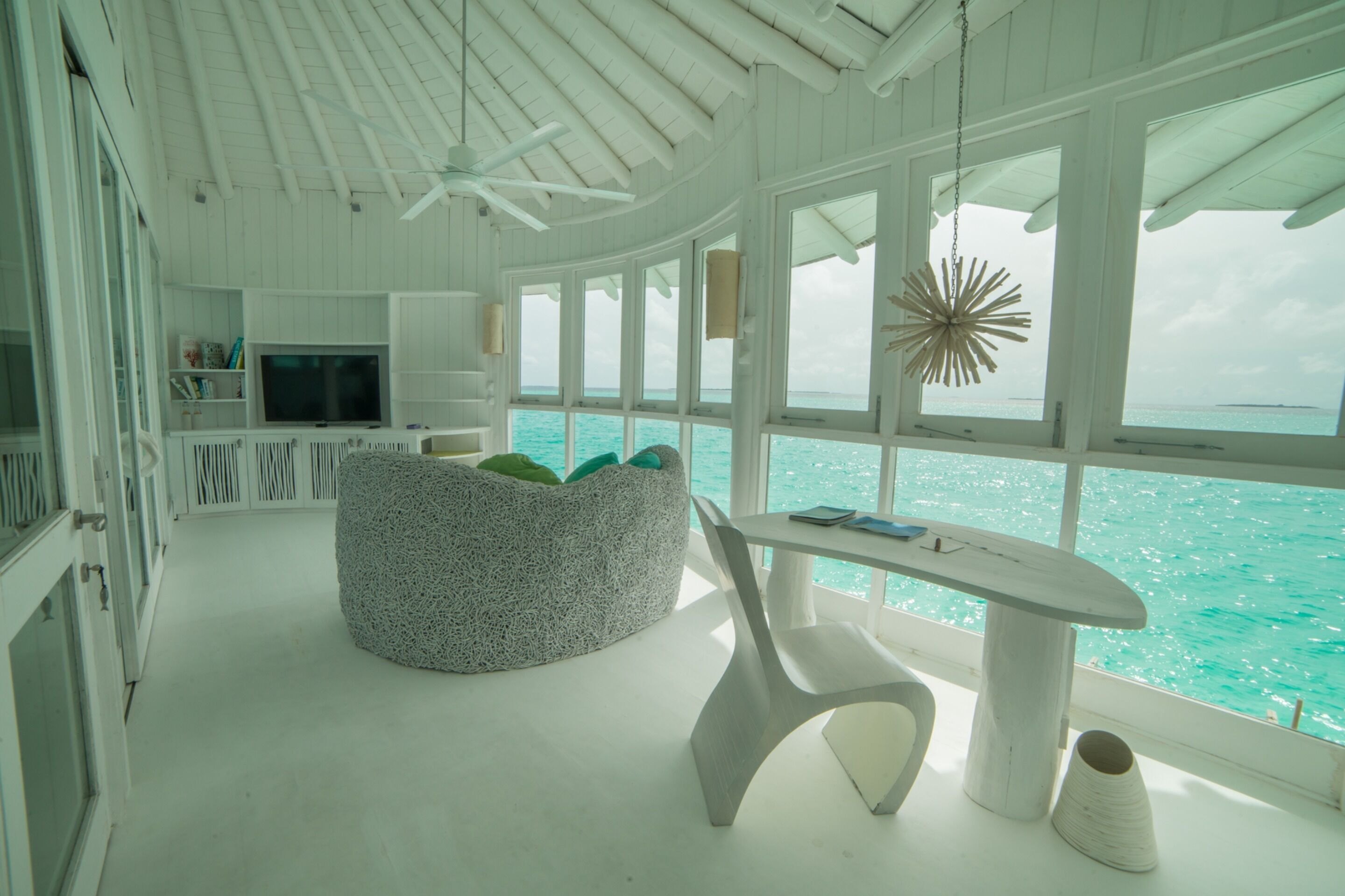 Una habitación moderna con decoración blanca, techo curvo, zona de estar circular, mesa con sillas, televisor y ventanas amplias con vista al océano.