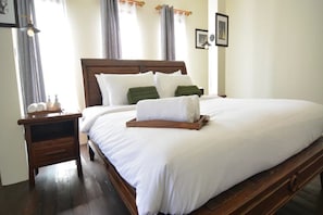 A neatly made bed with white linens and a wooden headboard, a wooden nightstand with a lamp, and a framed picture on the wall. - Nawa Saraan (Chiang Mai)