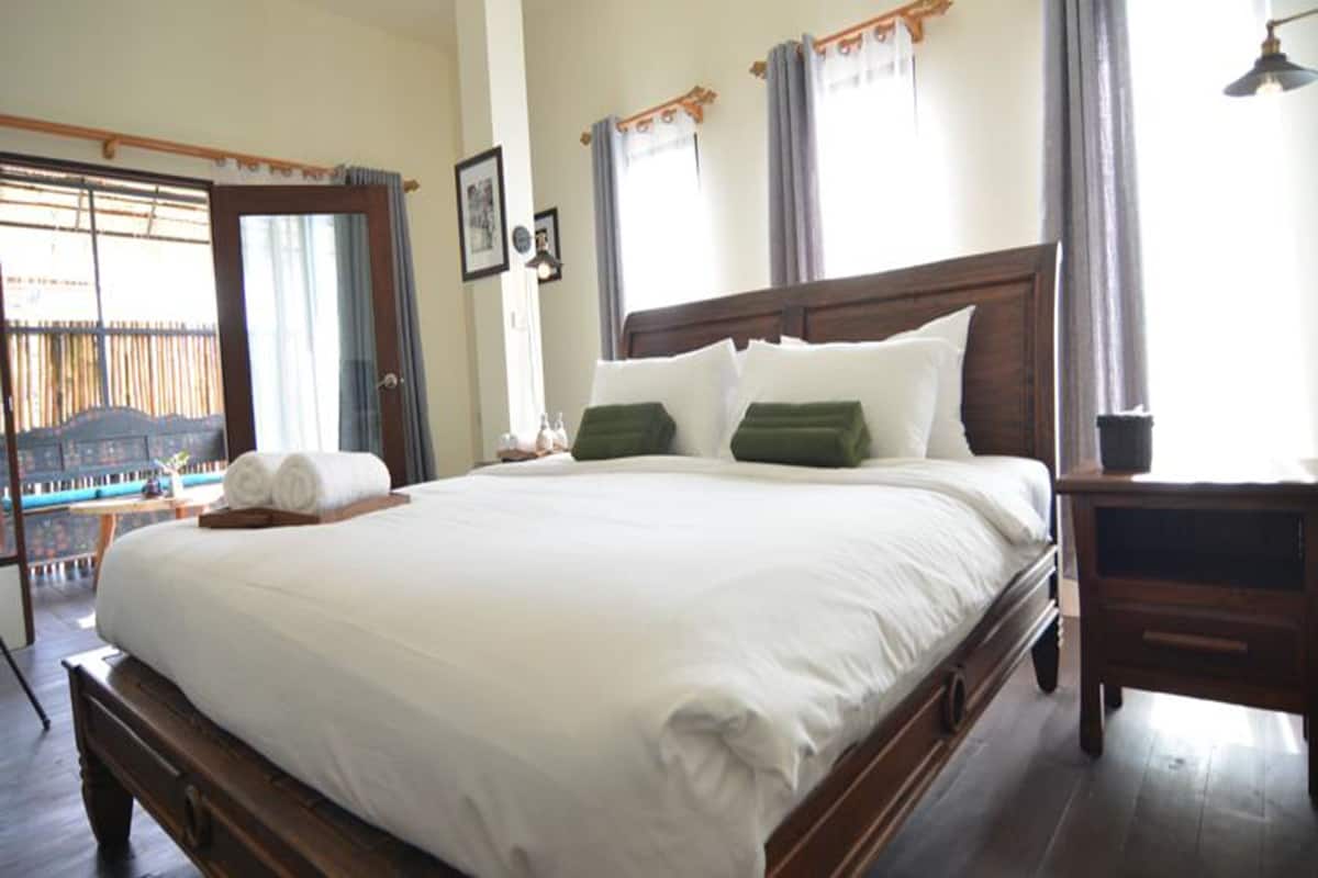 A neatly made bed with white linens, a wooden headboard, and green pillows. There is a wooden nightstand beside the bed, and a window with curtains to the right.