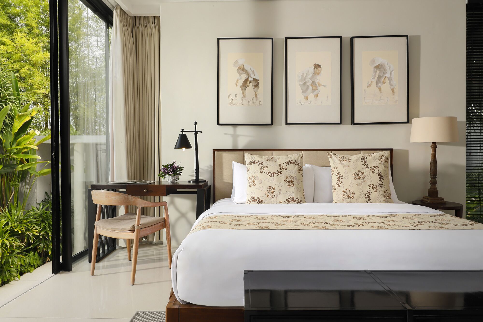 A hotel room with a bed, a desk, a chair, and a window with curtains. There are three framed pictures on the wall above the bed.
