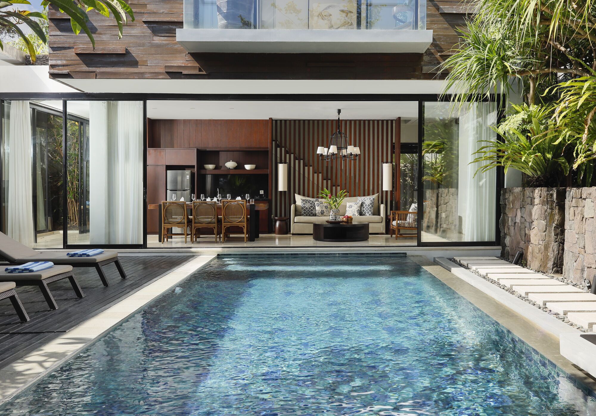 Une maison moderne dotée d’une piscine, d’un coin repas et d’une cuisine visible à travers de grandes portes vitrées.