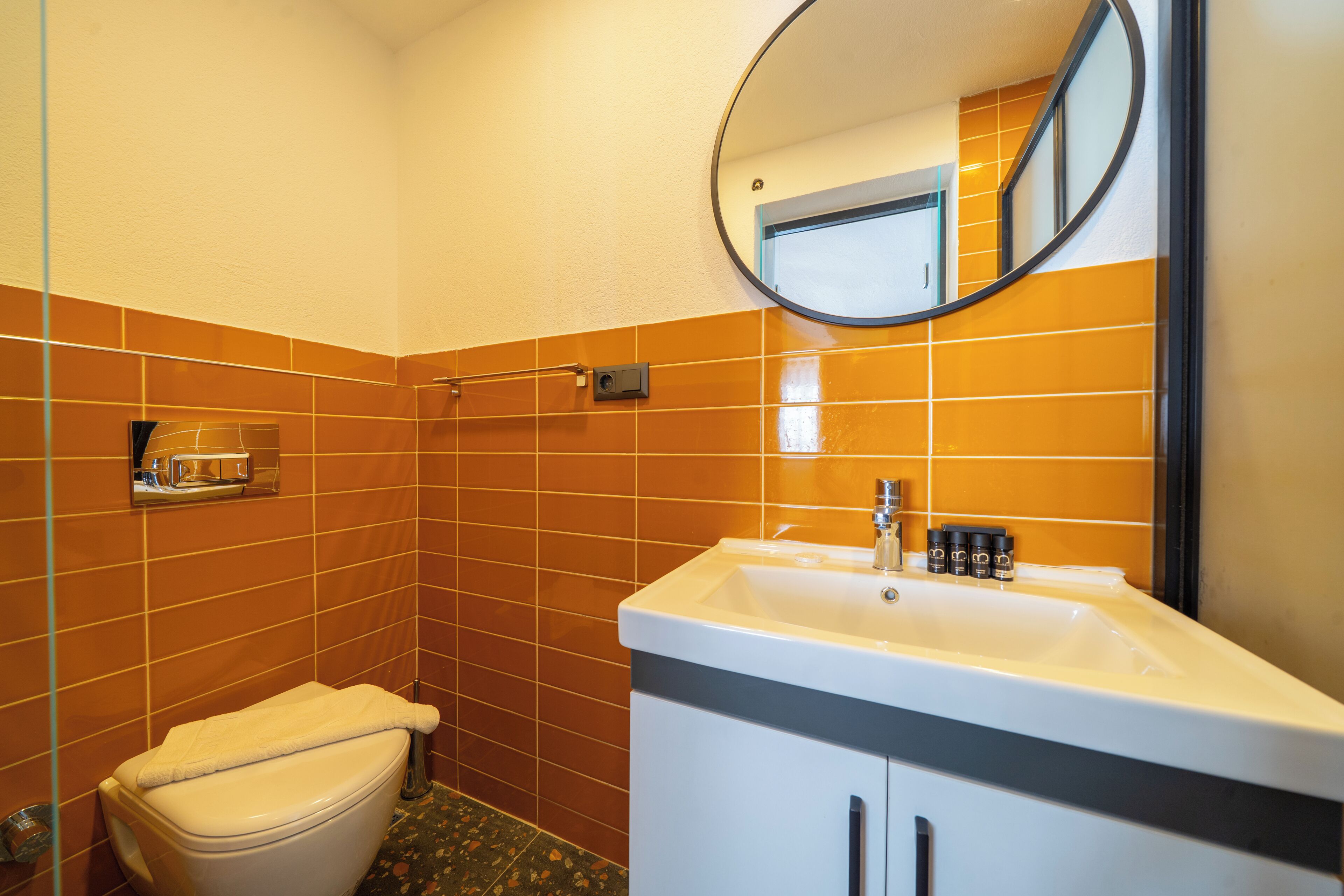 Un baño con inodoro, lavamanos, espejo y paredes con azulejos naranja.