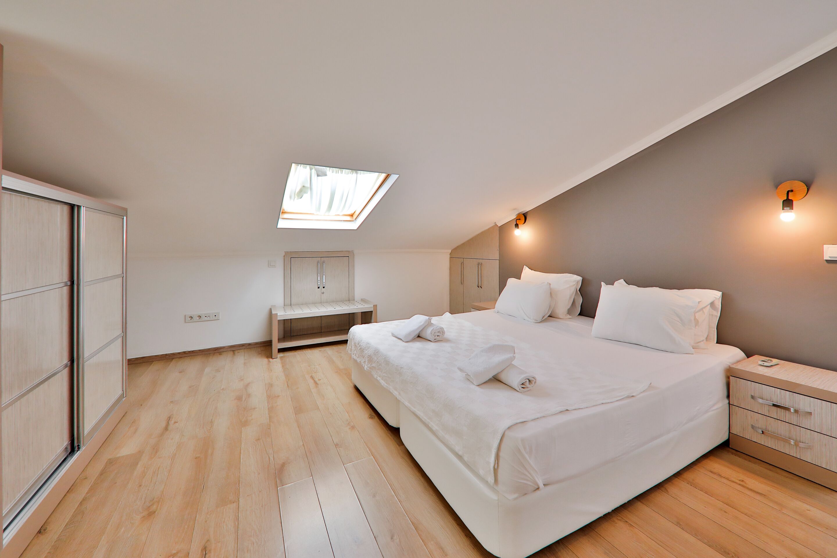 A bedroom with a skylight, a bed with white linens, a bedside table, and a wardrobe.