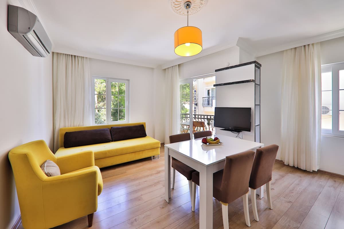 Una sala moderna con un sofá amarillo, una mesa de comedor con sillas, un televisor y un ventanal con cortinas.