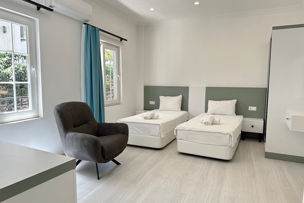 Una habitación de hotel moderna con dos camas individuales, un sillón gris, una mesita de noche pequeña y una ventana con cortinas.