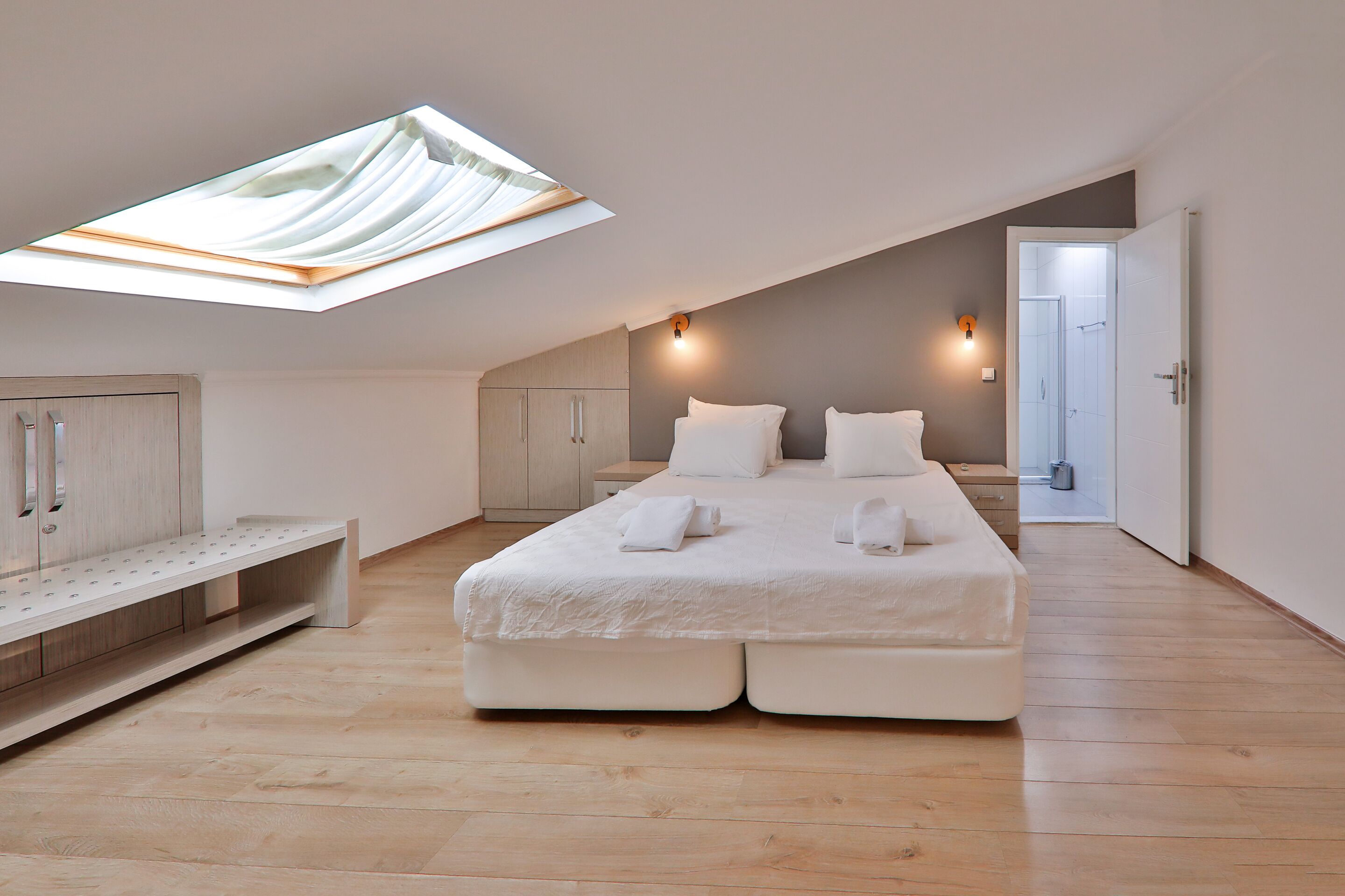 A modern bedroom with a skylight, a bed with white linens, a bench, and a wardrobe.