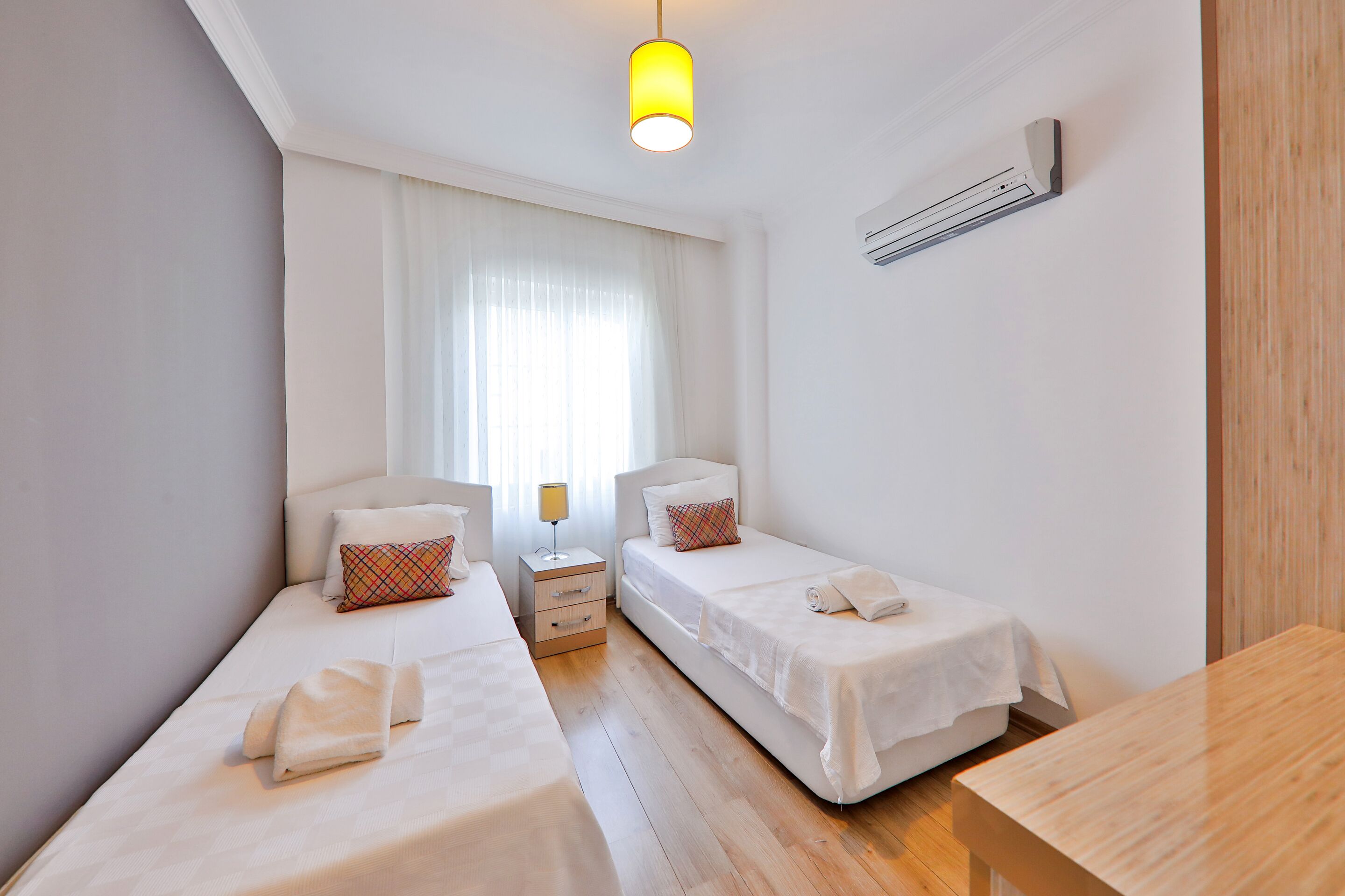 A clean, modern hotel room with two single beds, a wooden table, a bedside lamp, and a window with sheer curtains.