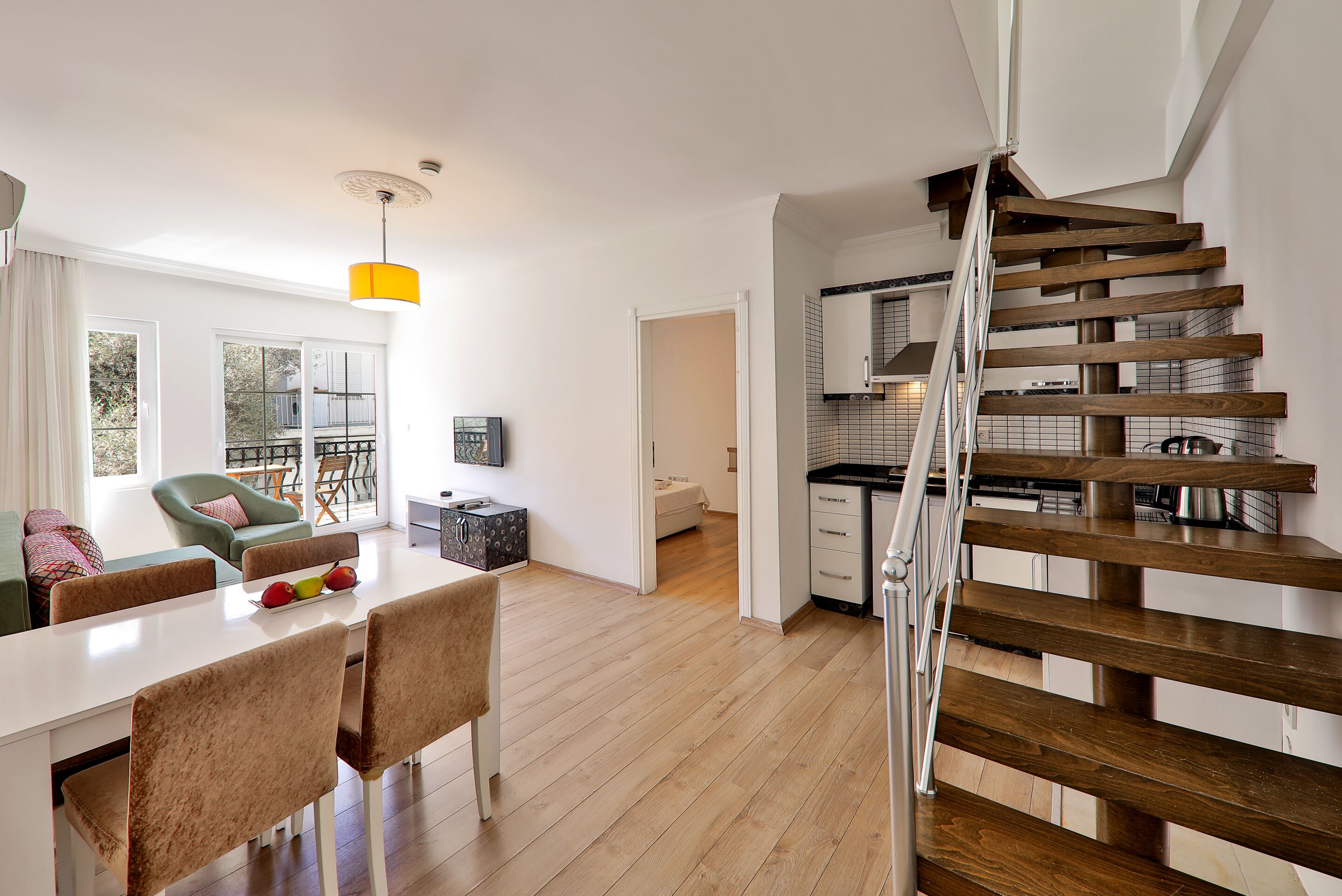 A modern apartment with a wooden staircase, a dining area with a white table and chairs, and a kitchen with white cabinets.