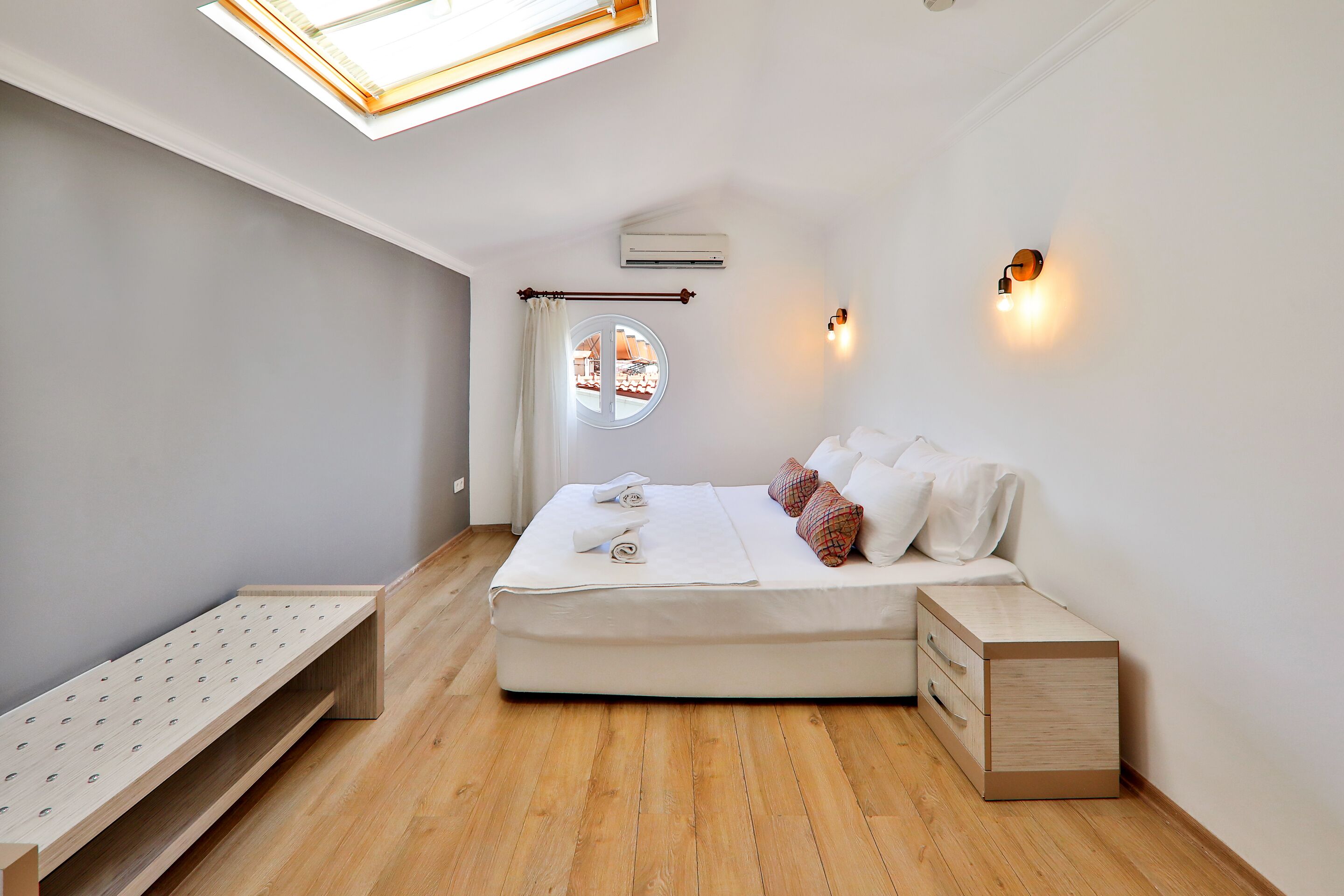 A modern bedroom with a skylight, a bed with white bedding, a bedside table, and a small wooden bench.