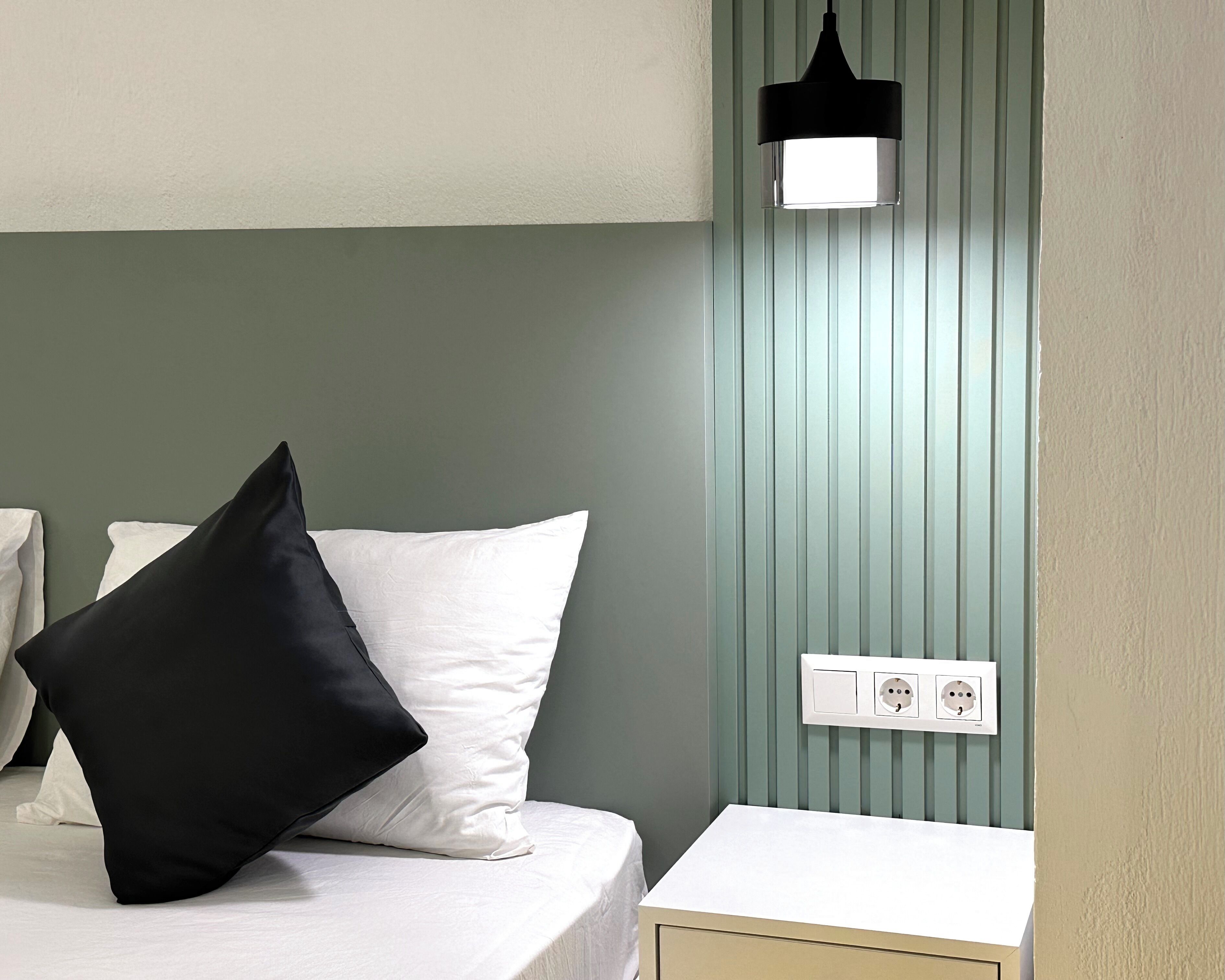 Een net opgemaakt bed met witte en zwarte kussens, een nachtkastje en een wandlamp.