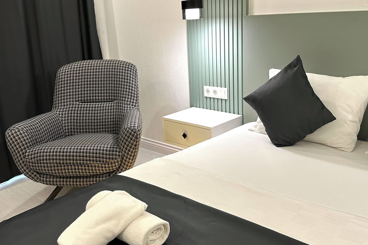 Habitación de hotel con un sillón a cuadros Príncipe de Gales, una cama con almohadas blancas y negras, una mesita de noche con una lámpara y una toalla blanca enrollada sobre la cama.