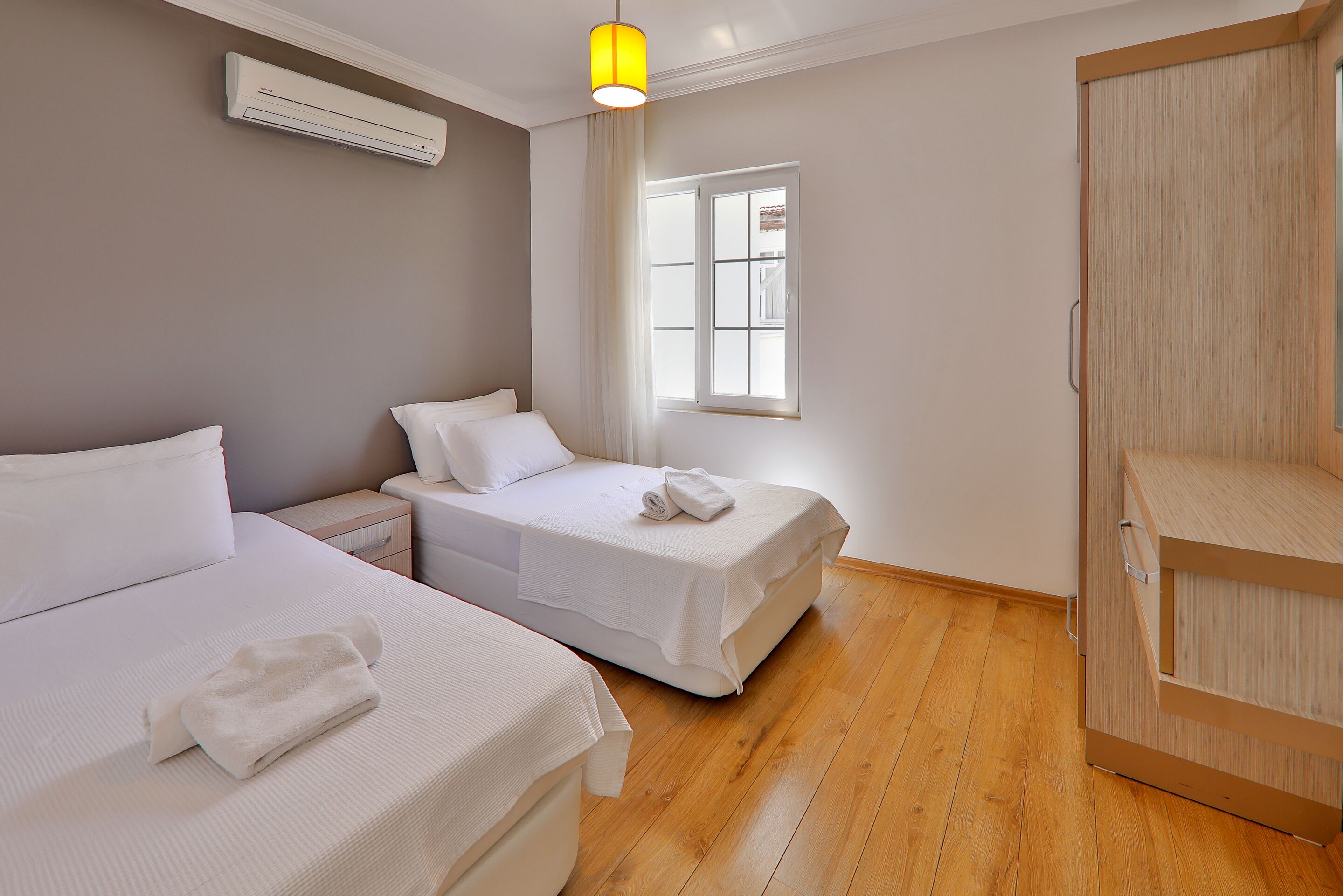 A hotel room with two beds, a wooden wardrobe, a window with curtains, and an air conditioning unit.