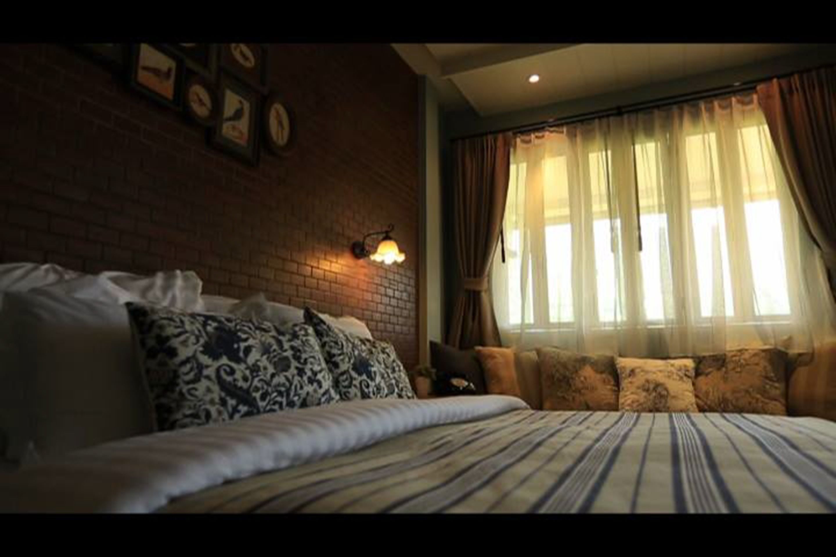 A bedroom with a brick wall, a bed with patterned pillows, a nightstand, and a window with curtains.