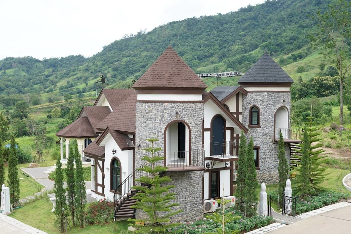 Une grande maison à étages, dotée d’une façade en pierre et de plusieurs toits à pignons, entourée d’une végétation luxuriante et de collines.