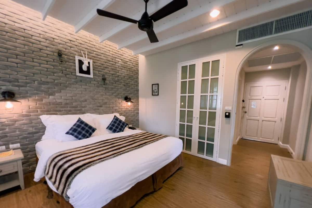 Une chambre avec un mur en briques, un lit avec une literie blanche, un ventilateur de plafond et un plancher en bois.