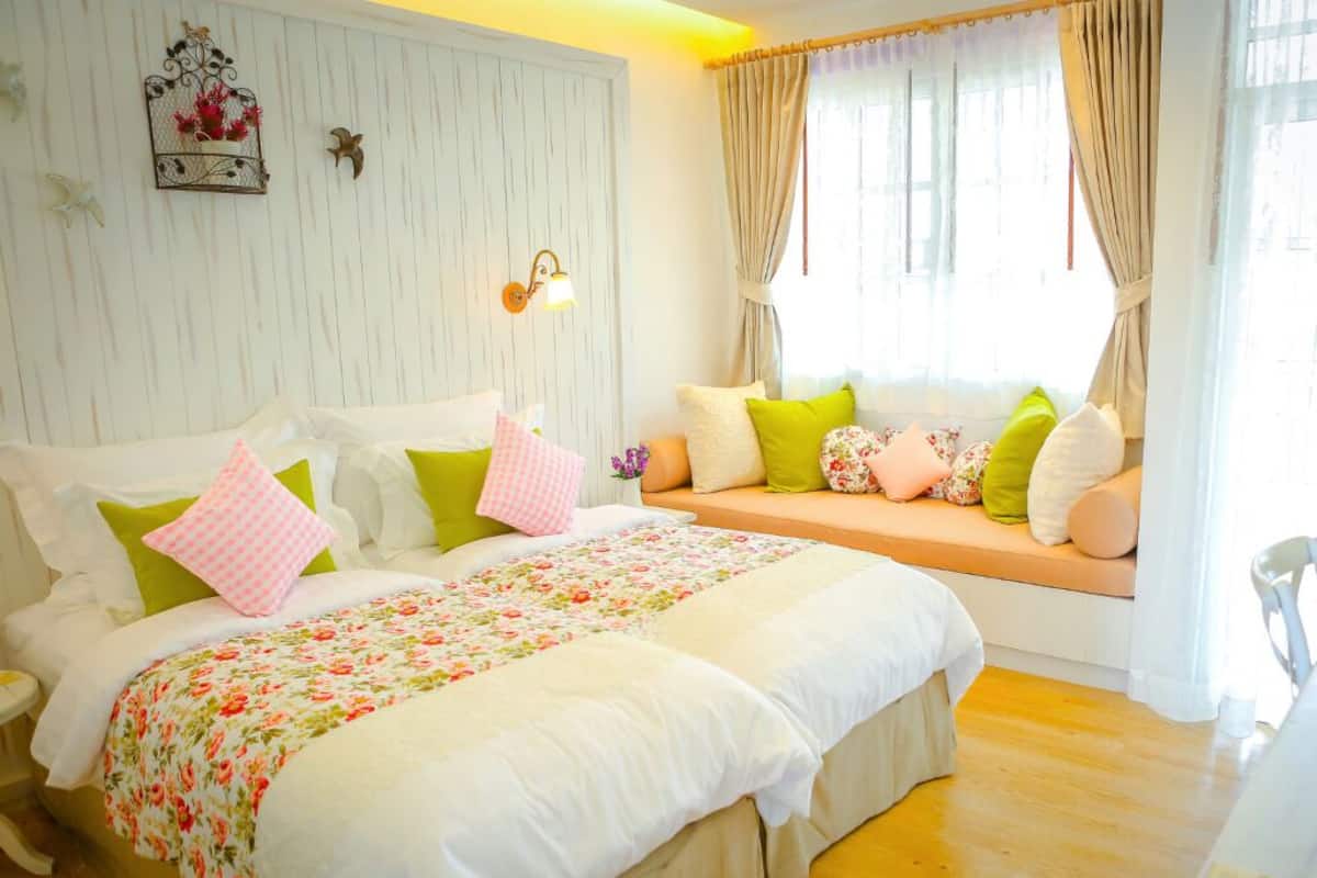 Une chambre avec un grand lit, un banc avec des coussins, une lampe fixée au mur et une fenêtre avec des rideaux.